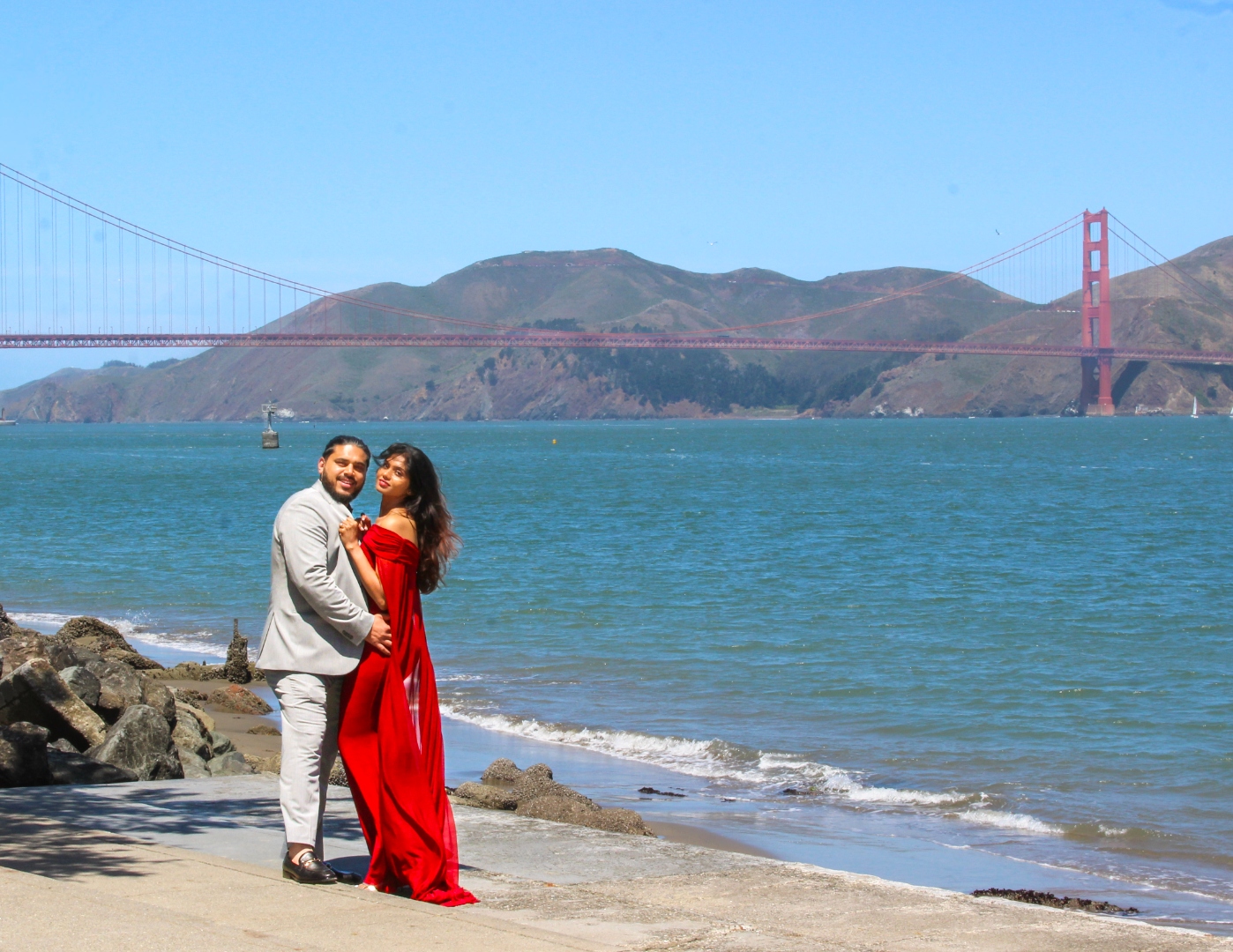 Prunella and Johnetan standing together on the shore with the Golden Gate Bridge tower rising behind them, in black and white